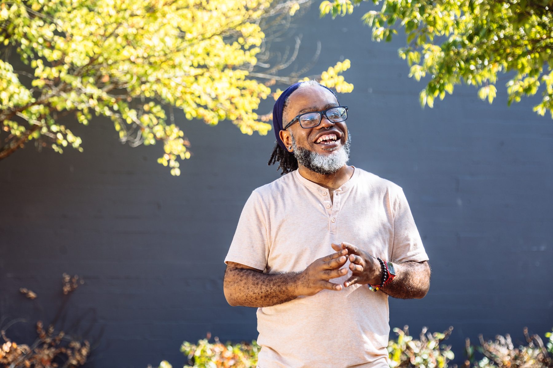 A Deaf man laughs mid-sign outdoors in dappled sunlight, his hands together in front of him.